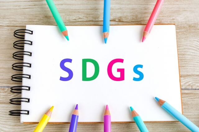 SDGsと書いてあるメモ帳の周りに8本の色鉛筆が円状に並んでいる