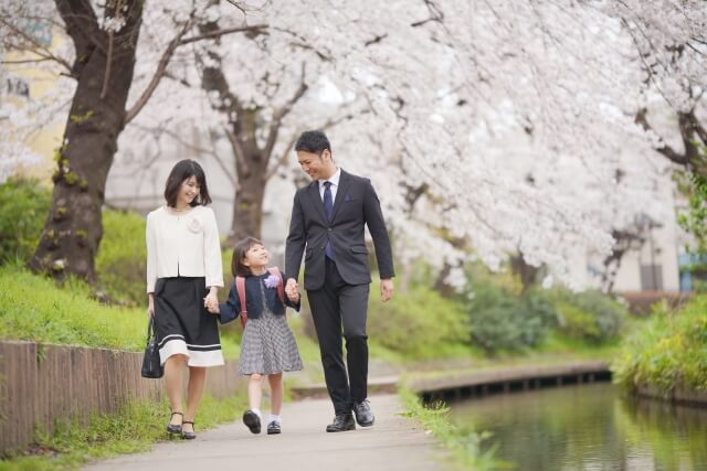 桜の木の下で子供と父親・母親が手をつないで歩いている画像