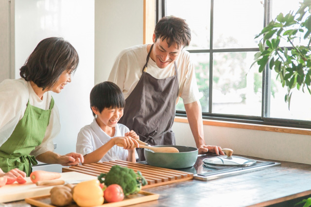 子供が料理をしている横で見守る両親の画像