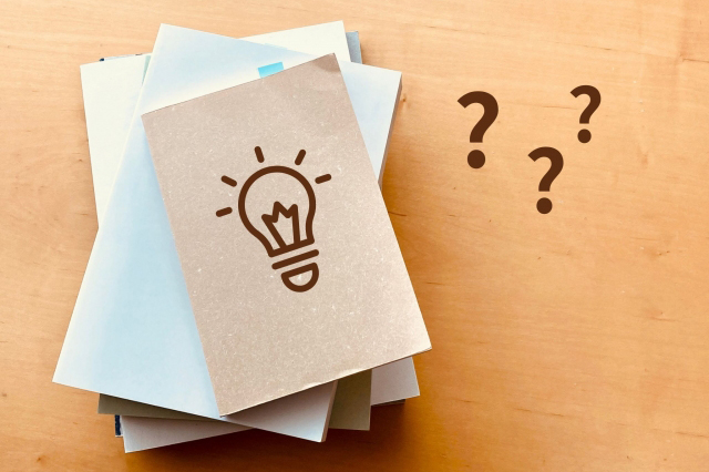 左側に積まれた本の表紙にひらめきマークが描かれ、右側に?マークが描かれている画像
