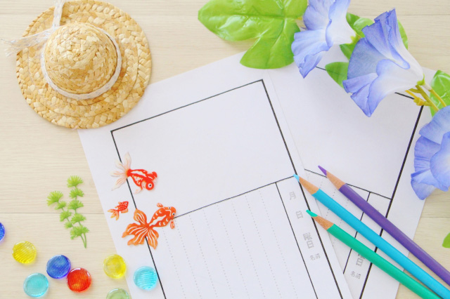 白紙の絵日記の上に、朝顔の花、麦わら帽子、色鉛筆、おはじき、金魚の置物が並べて置かれている画像