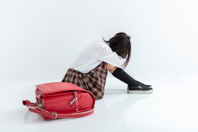 膝を抱えてうつむく子供とその傍らにランドセルが置いてある画像